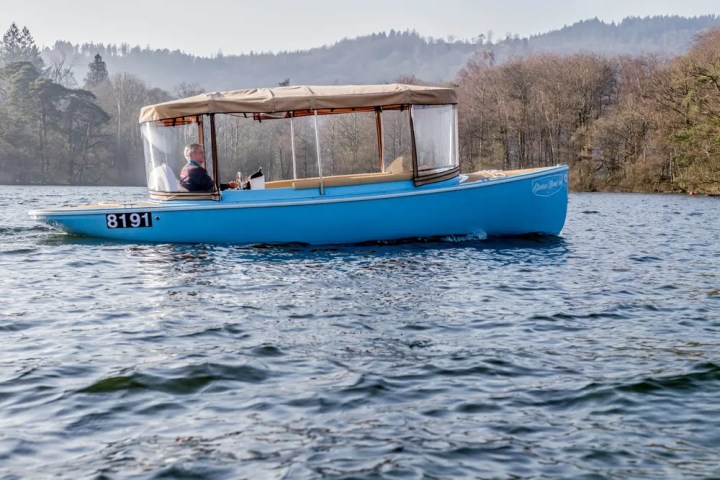 Blue boat with canopy on a lake, person steering, forest and hills in background.