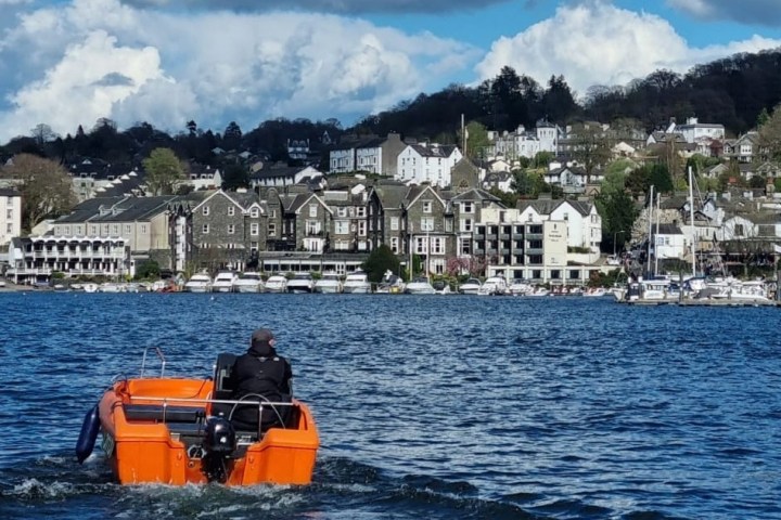 Person in orange boat on lake approaching a coastal town with hills and clouds in the background.