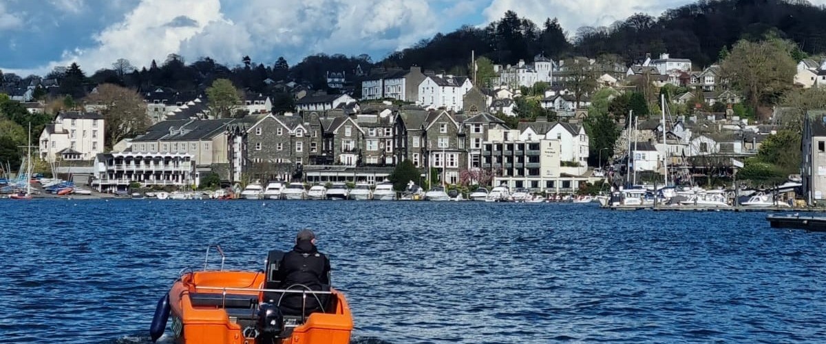 Person in orange boat on lake approaching a coastal town with hills and clouds in the background.