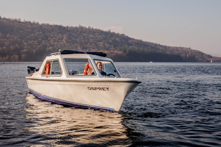 A small white boat named Osprey with a person inside, cruising on a calm lake on a clear day.