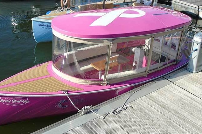 Pink electric boat with a white ribbon on top, docked at a marina with wooden decking.