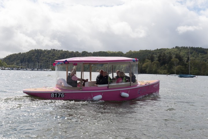 Pink boat with people on a lake, surrounded by hills and trees.